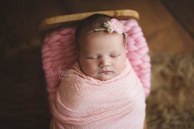 A sleepy newborn girl in pink with headband on a wood floor backdrop 