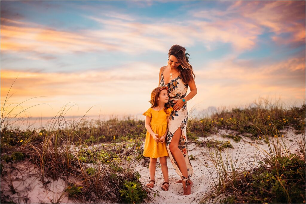 Clearwater Beach, FL Family Photography. Mother and daughter lovingly embrace at sunset on the sand dunes at Clearwater beach during golden hour full of radiant sunset colors